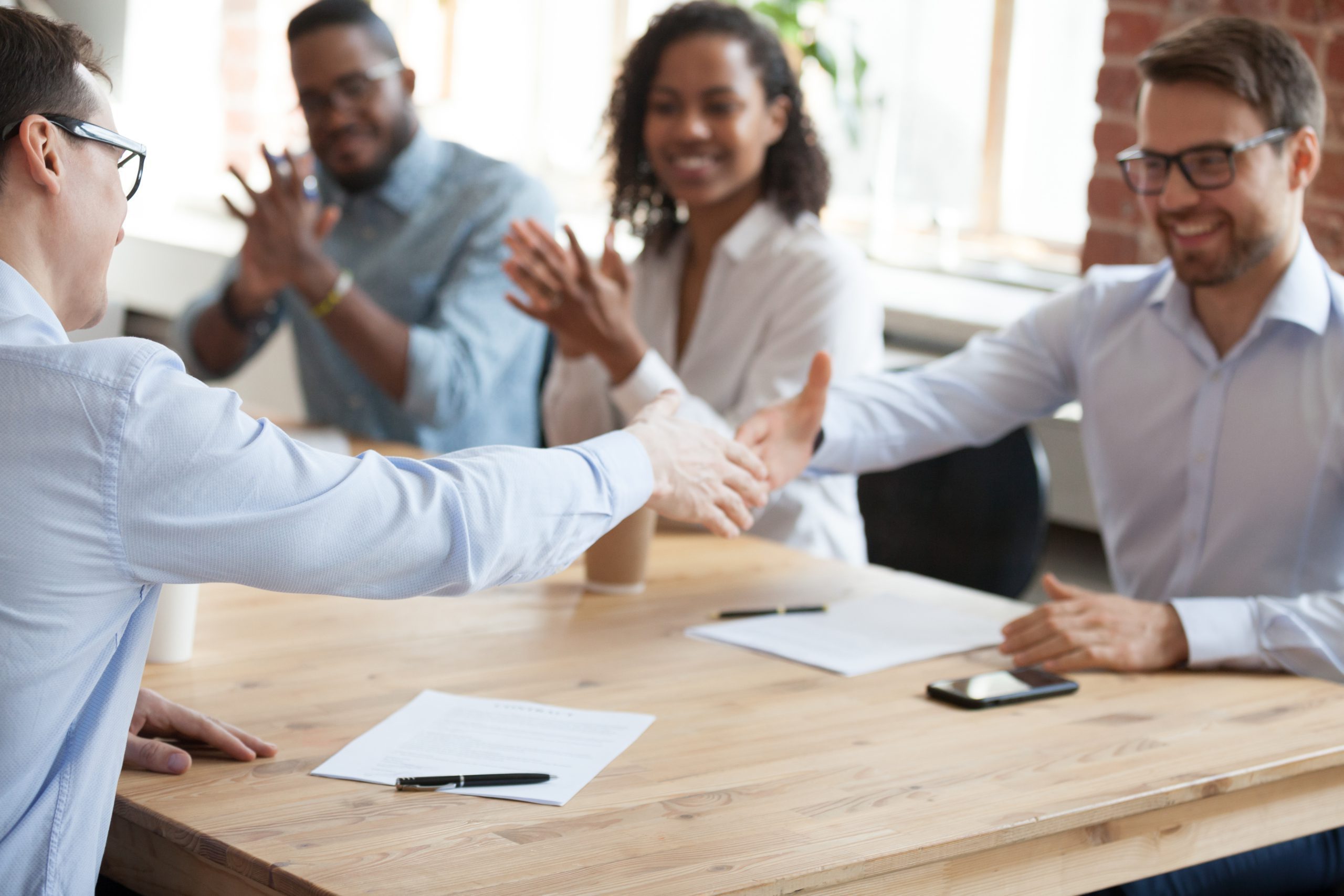 Two colleagues shaking hands across a table while others smile and clap in the background during a meeting.