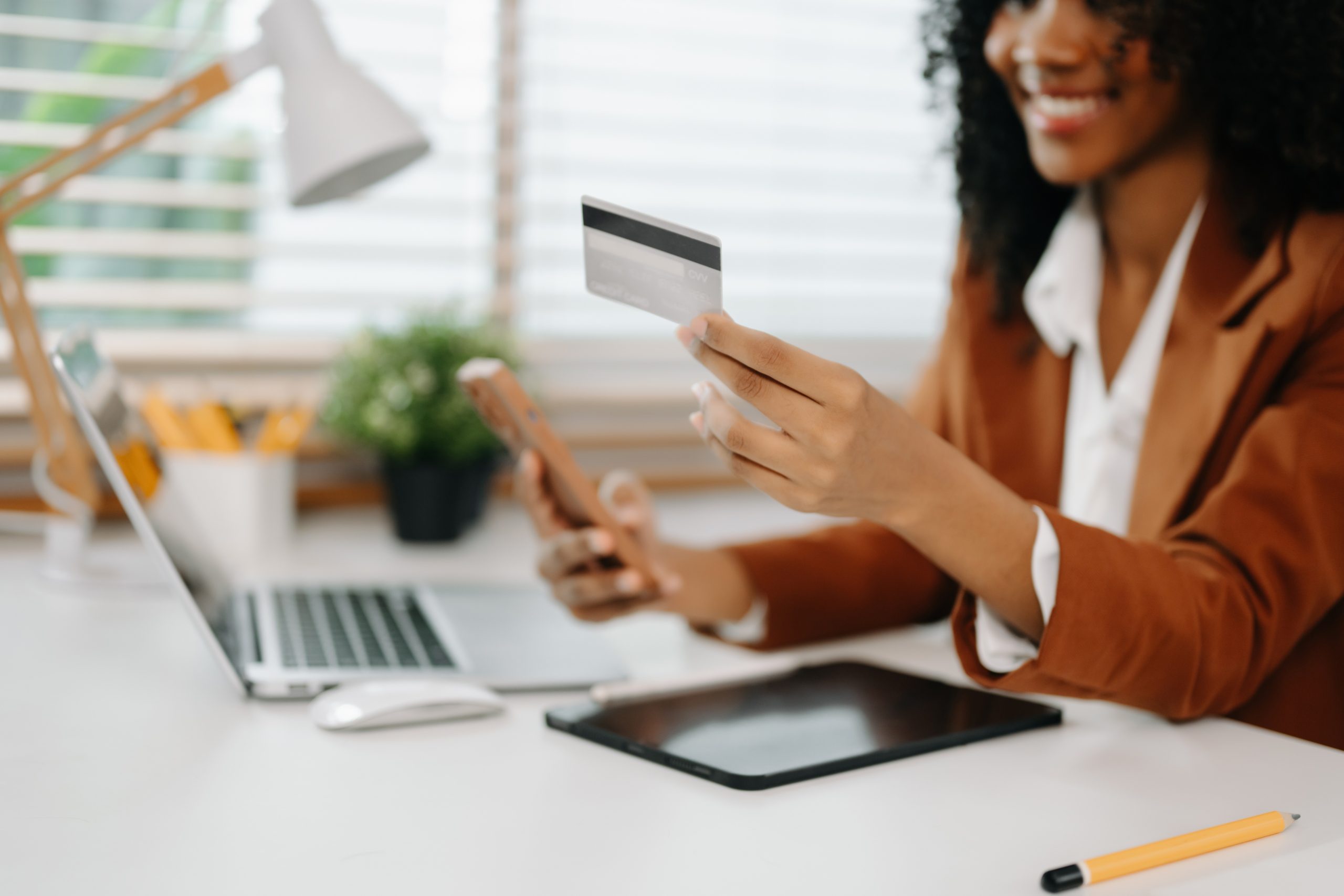 Woman holding a credit card and smartphone while sitting at a desk with a laptop, making an online purchase.