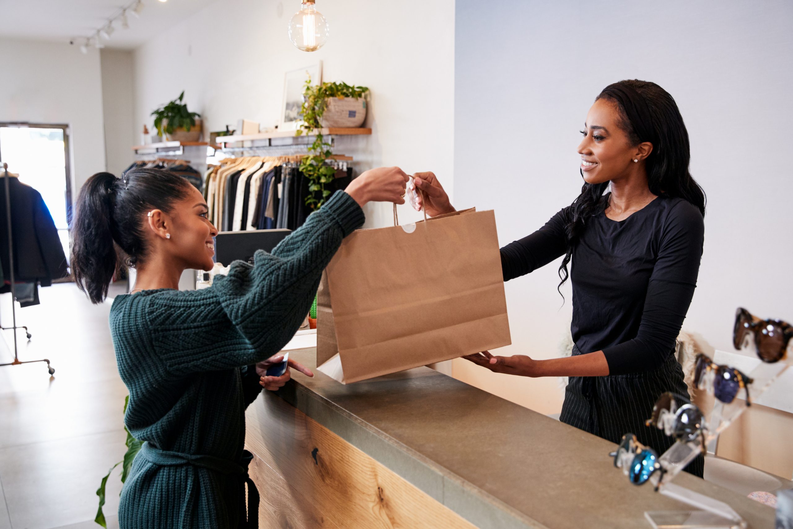 Retail associate handing a shopping bag to a customer at a boutique checkout counter, with clothing displays in the background.